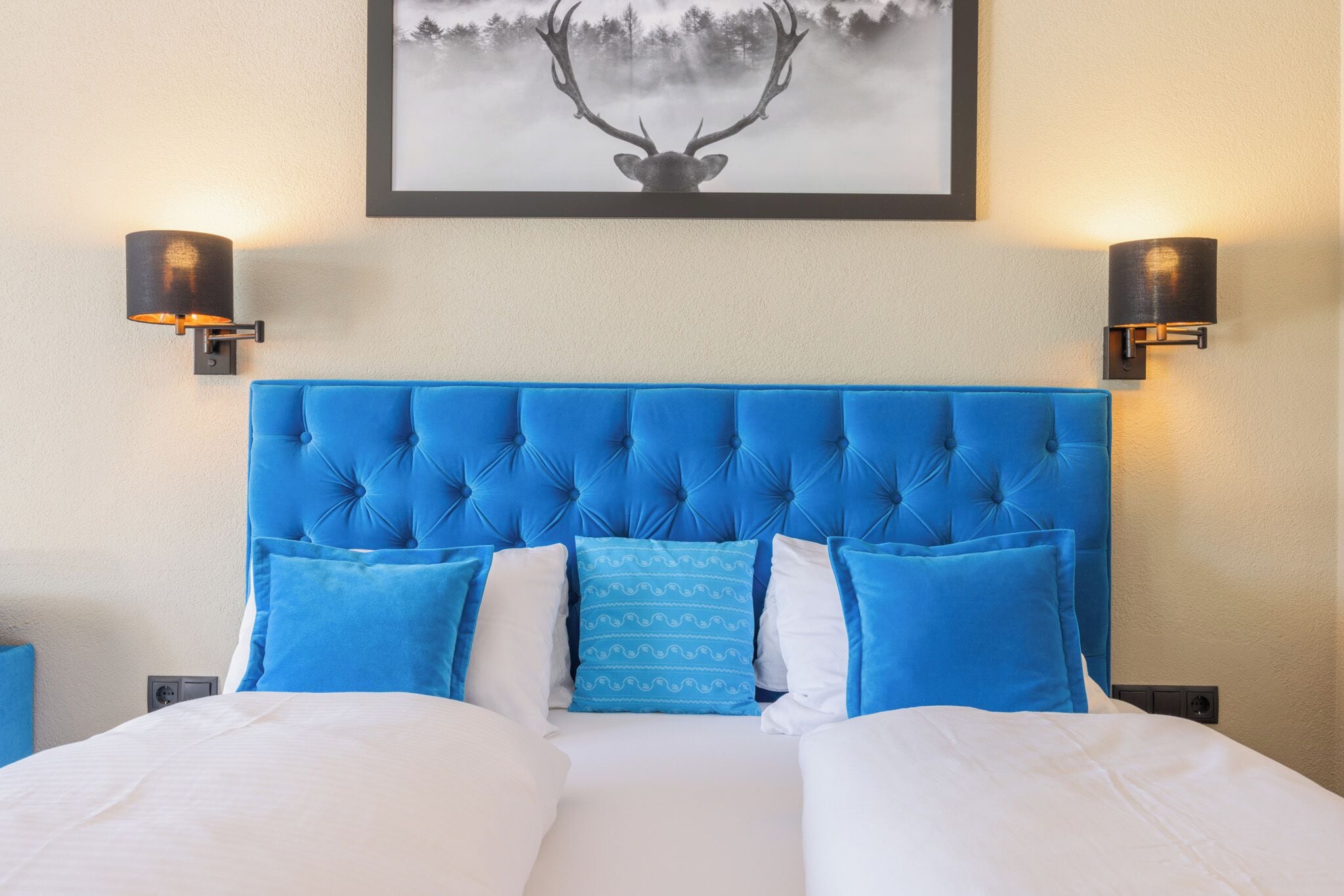 Ein Hotelzimmer mit blauem, gesteptem Kopfteil, weißer Bettwäsche und einem Hirschbild über dem Bett.