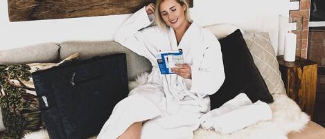 Una persona con bata blanca sentada en un sofá con un libro, rodeada de almohadones y una alfombra blanca mullida.