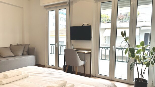 A hotel room with a large bed, a desk, a chair, a TV, and a balcony with a potted plant.