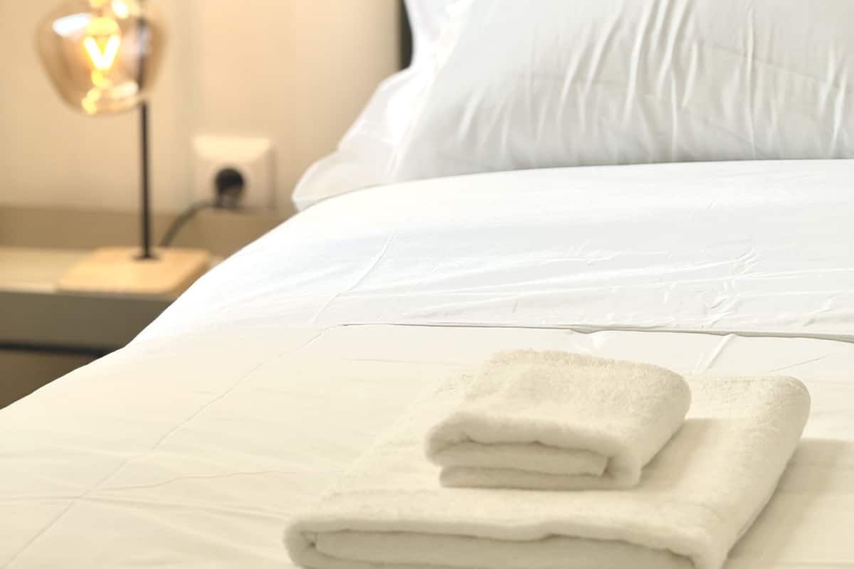 Ein ordentlich bezogenes Bett mit weißen Leinen und einem gefalteten Handtuch daneben.
