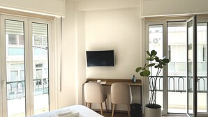 A hotel room with a bed, a desk, two chairs, a TV, and a balcony with a potted plant.