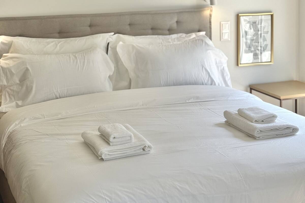 Ein Hotelzimmer mit einem großen Bett, weißen Bettwäsche, einer Kopfstütze, einer Klimaanlage, einem Nachttisch und einem gerahmten Bild an der Wand.
