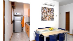 Une cuisine compacte équipée d’un réfrigérateur, d’un four à micro-ondes et d’une serviette orange accrochée à la porte. Une table à manger avec des chaises bleues et un panier de fruits se trouve au centre de la pièce.