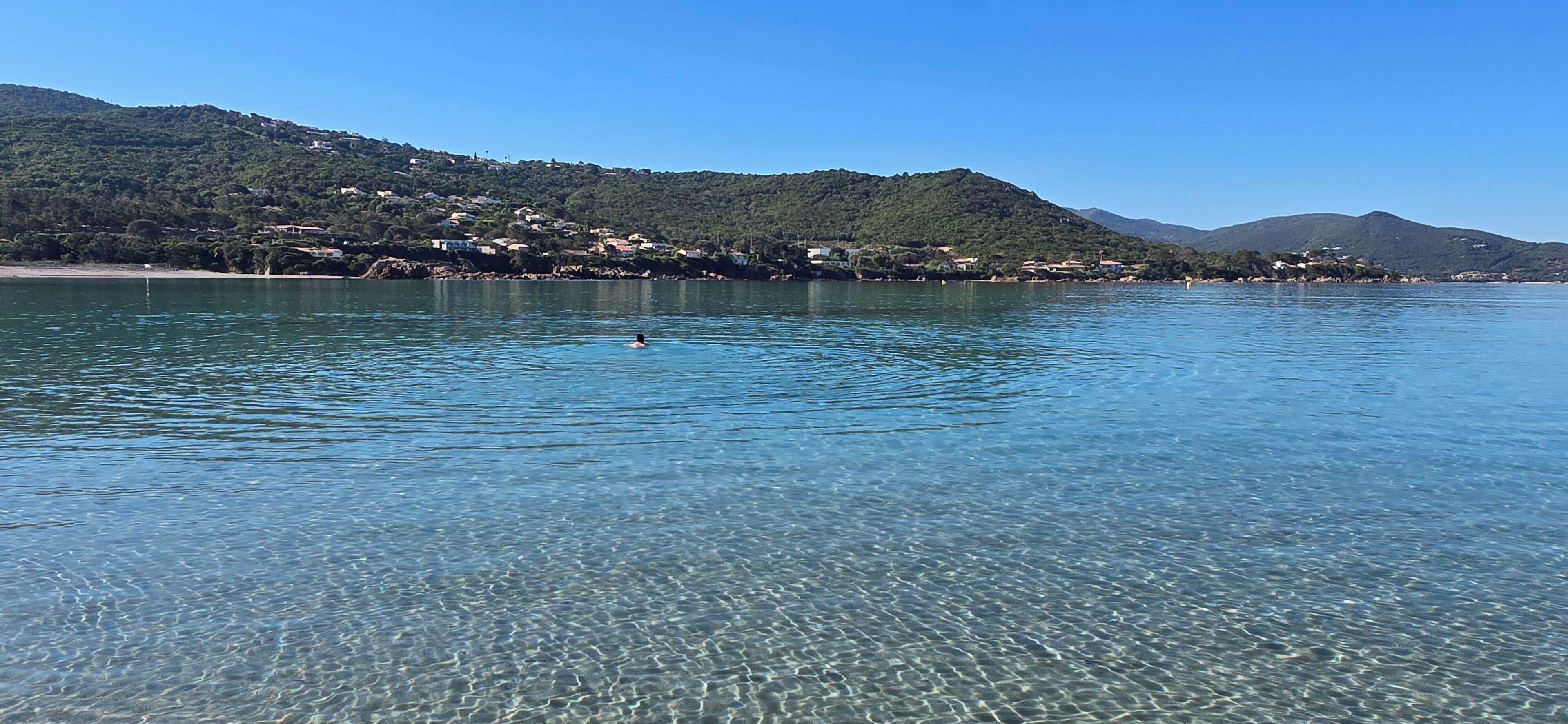 A clear lake with a swimmer in the water, surrounded by hills and buildings.