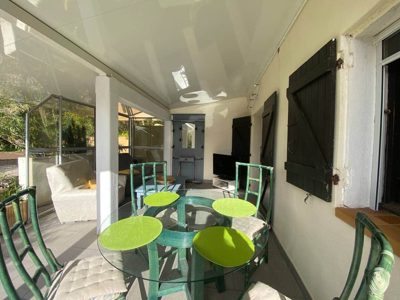 Um pátio externo com mesa e cadeiras de vidro, um sofá branco e uma televisão em um suporte.