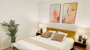 Un lit bien fait avec une tête de lit en bois, deux tables de chevet avec des lampes, une grande plante en pot et des œuvres d’art abstraites accrochées au mur.
