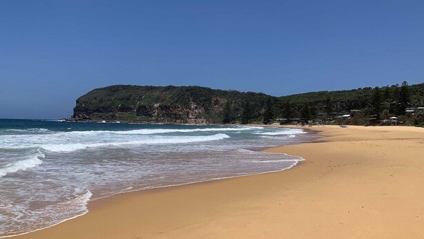 A wide sandy beach with gentle waves, a forested cliff, and a clear blue sky. - Retro Beach House by HolidayCo. (MACMASTERS BEACH)