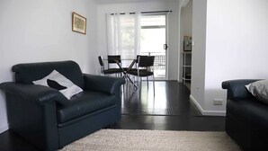 A modern living room with a black leather sofa, a dining area with a table and chairs, and a sliding glass door leading to a balcony. - Retro Beach House by HolidayCo. (MACMASTERS BEACH)