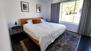 A bedroom with a large bed, a small bedside table, two framed pictures on the wall, and a window with curtains. - Retro Beach House by HolidayCo. (MACMASTERS BEACH)
