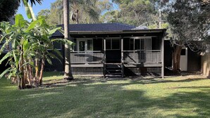 A two-story house with a porch, surrounded by lush greenery and trees. - Retro Beach House by HolidayCo. (MACMASTERS BEACH)