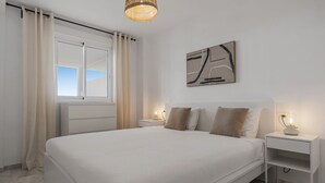A bedroom with a large bed, a window with curtains, a bedside table with a lamp, and a wall-mounted abstract art piece.