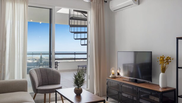 A modern living room with a large TV, a comfortable sofa, a dining table, and a balcony with a view of the sea.
