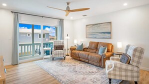 A spacious living room with a large window, a ceiling fan, a brown leather sofa, two plaid armchairs, a patterned rug, and a painting on the wall. - 4819B Stunning Townhouse - 2 bed/2.5 bath Pet Friendly! (Panama City Beach)
