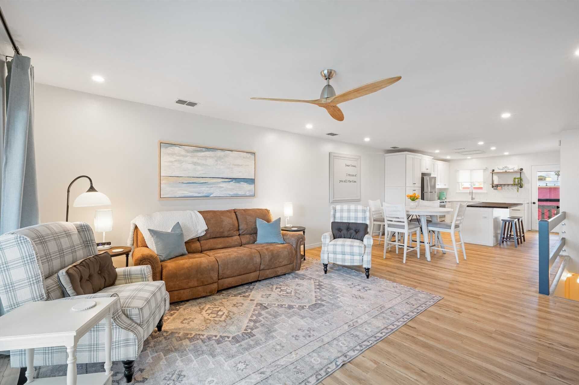 A modern living room with a brown sofa, a white coffee table, and a patterned rug. There is a dining area with a white table and chairs, and a kitchen in the background.