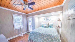 A bedroom with a wooden ceiling, a bed with a blue patterned bedspread, a ceiling fan, a window with blinds, and a door.