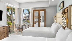 Ein Hotelzimmer mit zwei Betten, einem Schreibtisch und einem Stuhl. Durch die Glastür ist ein Blick auf Palmen und einen Pool.