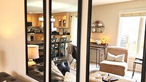 A room with a large mirror reflecting a dining area, a sofa, and a window with a view. - Everchanging Seasons: Charming Retreat + Cottage • Hot Tub • Sauna • Game Haven (Burnsville)