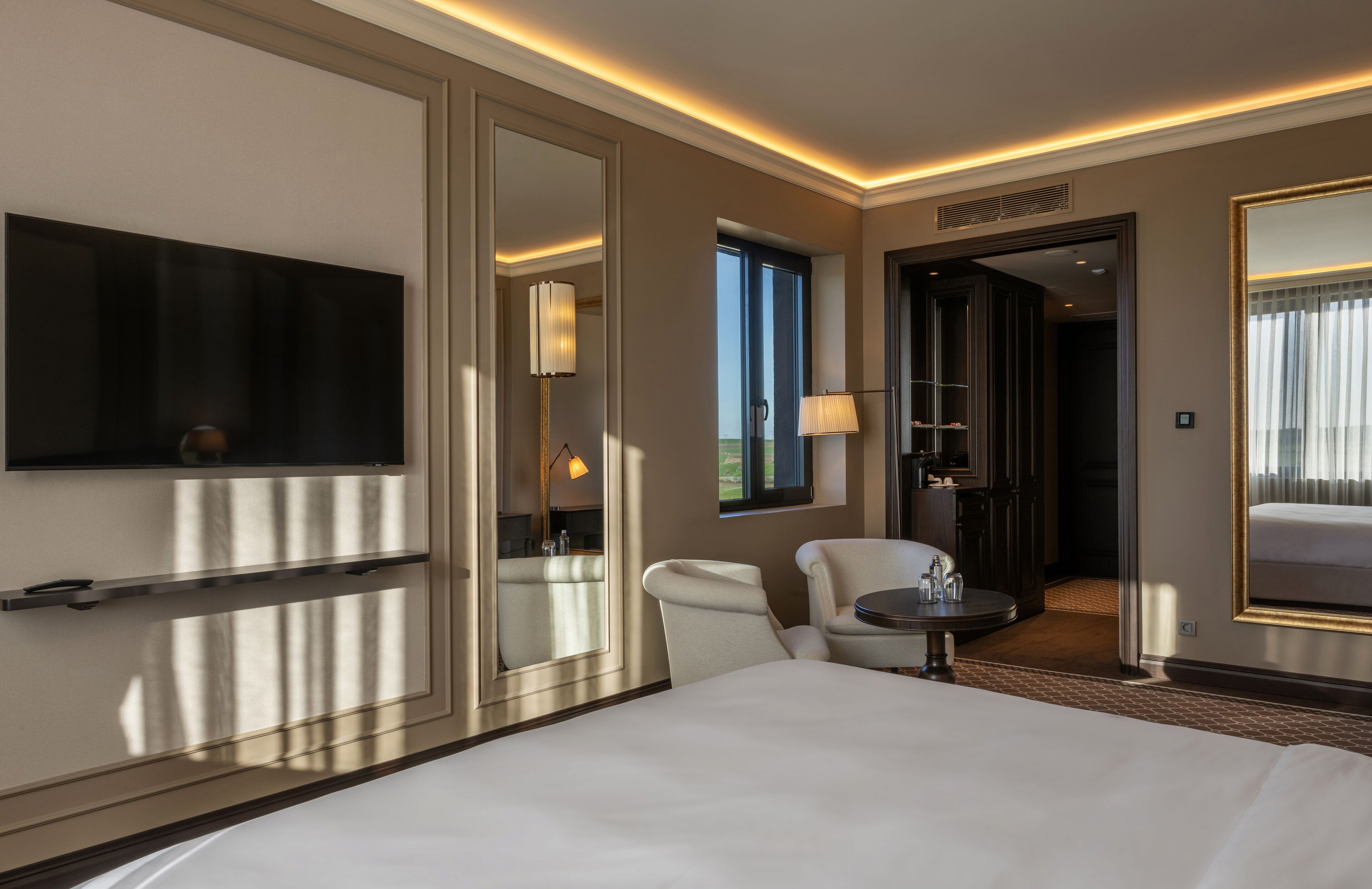 Una habitación de hotel con una cama grande, un televisor de pantalla plana, una zona de estar con una silla y una mesita pequeña, y un espejo grande.