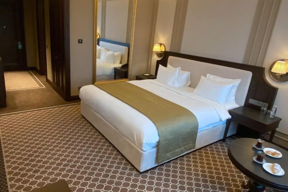 Une chambre d’hôtel avec un grand lit, une table de chevet avec une lampe, une table ronde avec des assiettes et des tasses, et un sol à motifs.