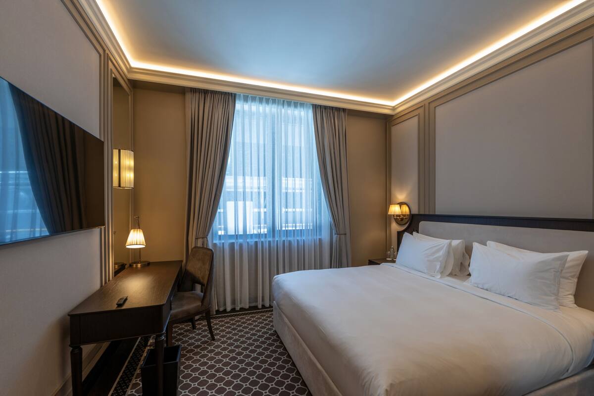 Une chambre d’hôtel avec un grand lit, un bureau, une chaise, une lampe et une fenêtre avec des rideaux.