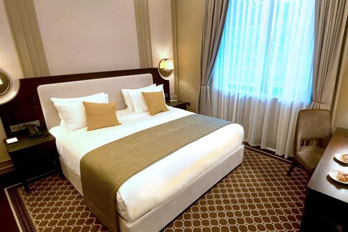 A hotel room with a large bed, a bedside table with a lamp, a chair, and a view of the outdoors through the window.