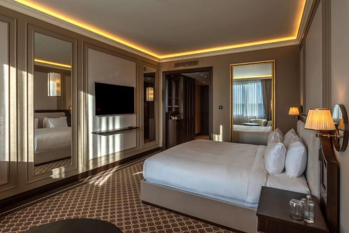 A hotel room with a large bed, a flat-screen TV, a bedside table with a lamp, and a mirror.