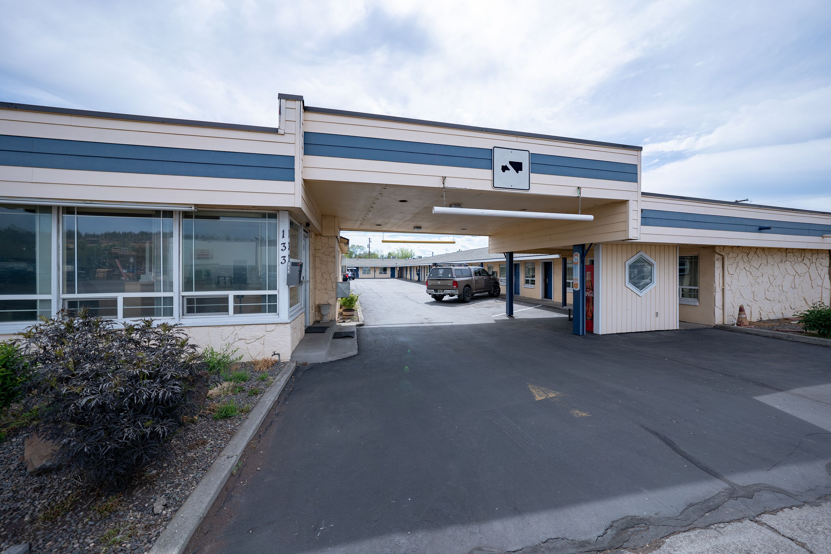 Un motel con área de estacionamiento techada, un ventanal grande a la izquierda y un pequeño edificio con ventanilla a la derecha.