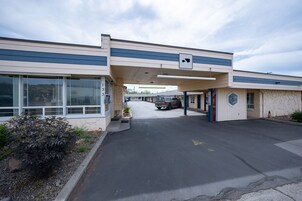Un motel con área de estacionamiento techada, un ventanal grande a la izquierda y un pequeño edificio con ventanilla a la derecha.