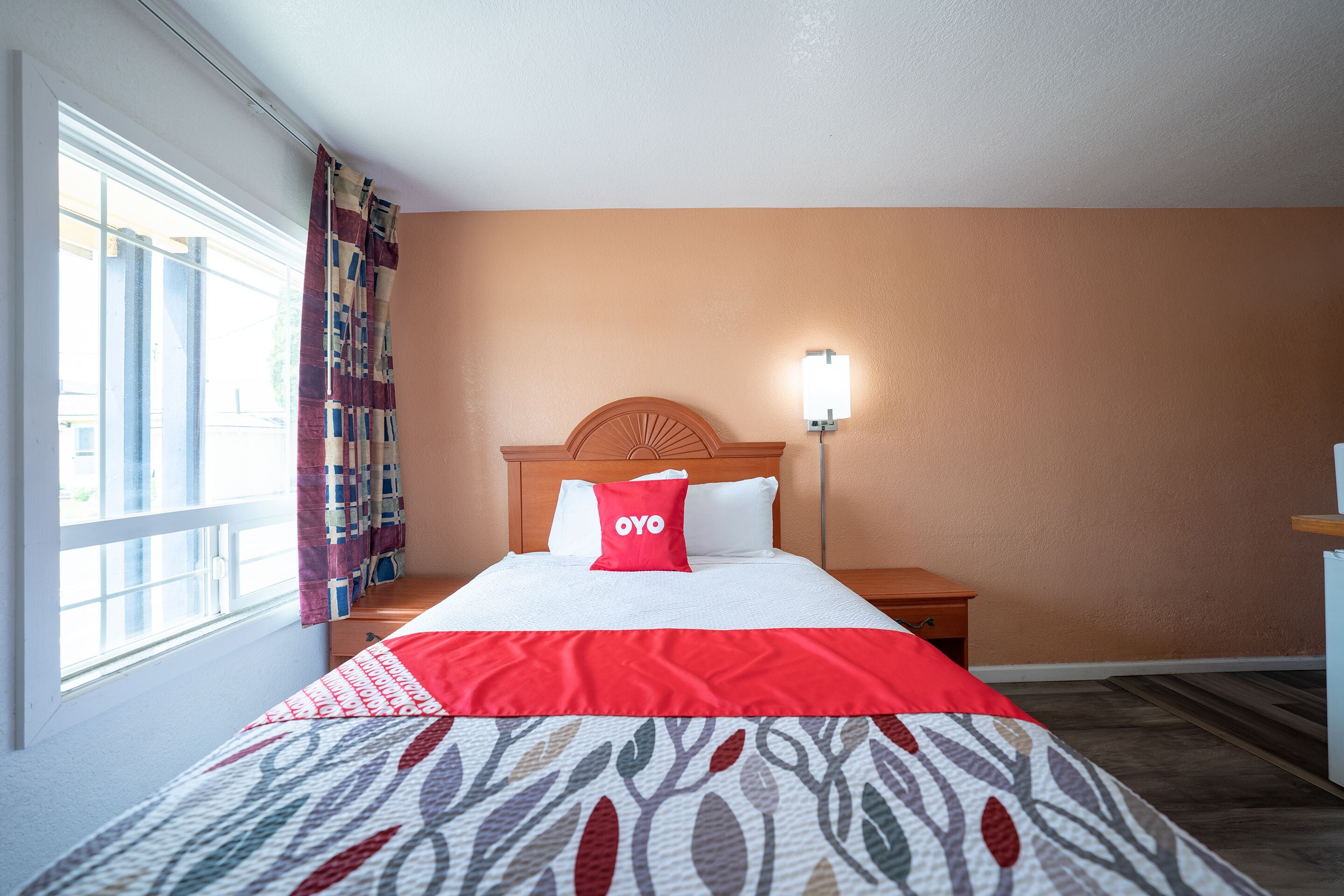 Habitación de hotel con cama, mesita de noche, lámpara de pared y ventana con cortinas.