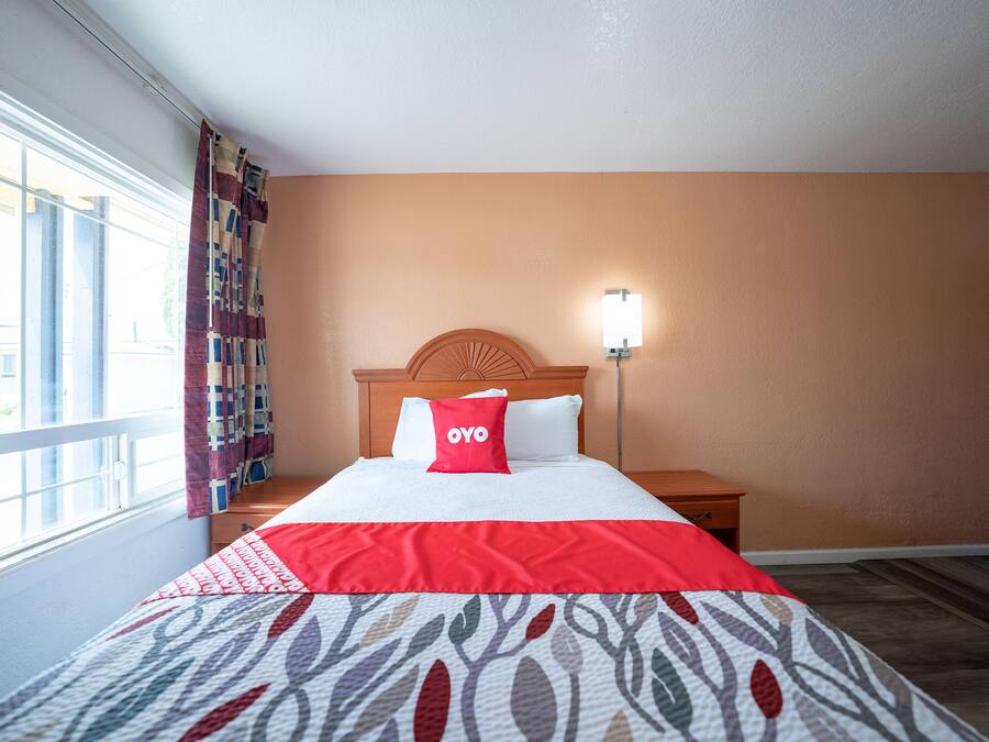Habitación de hotel con cama, mesita de noche, lámpara de pared y ventana con cortinas.