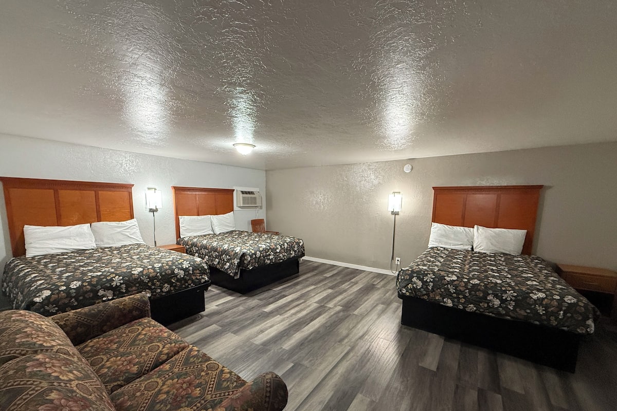 A hotel room with three beds, a wooden headboard, a standing lamp, and a patterned sofa.