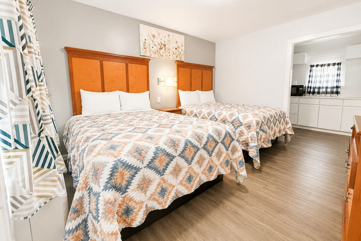 A hotel room with two beds, a wooden headboard, a nightstand, a wall-mounted light, and a kitchenette.