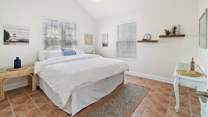 Une chambre avec un grand lit, deux fenêtres équipées de stores, une petite table avec une lampe et des œuvres d’art accrochées au mur.