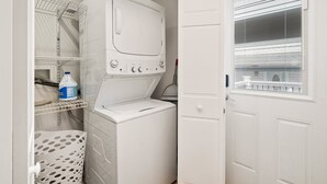 Une buanderie équipée d’un lave-linge et d’un sèche-linge superposés, d’un étendoir et d’une étagère de rangement où se trouve une bouteille de lessive.