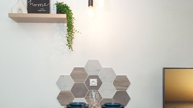 Een moderne eetruimte met een houten tafel, twee zwarte stoelen, een betegelde achterwand in een honingraatmotief, een klein plankje met een plant en een aan de muur bevestigde televisie.