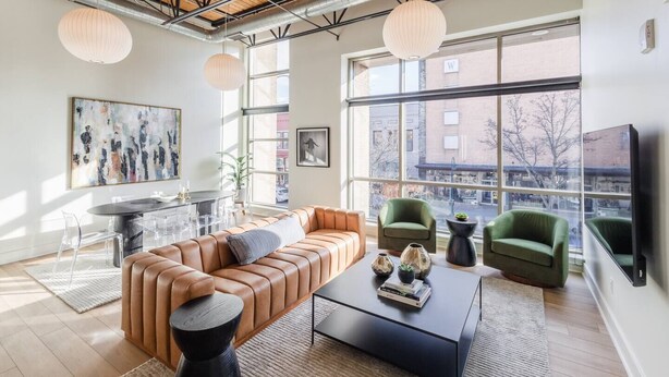 A modern living room with a leather sofa, green armchairs, a black coffee table, and large windows offering a city view. - Modern Luxury in Downtown TC (Traverse City)