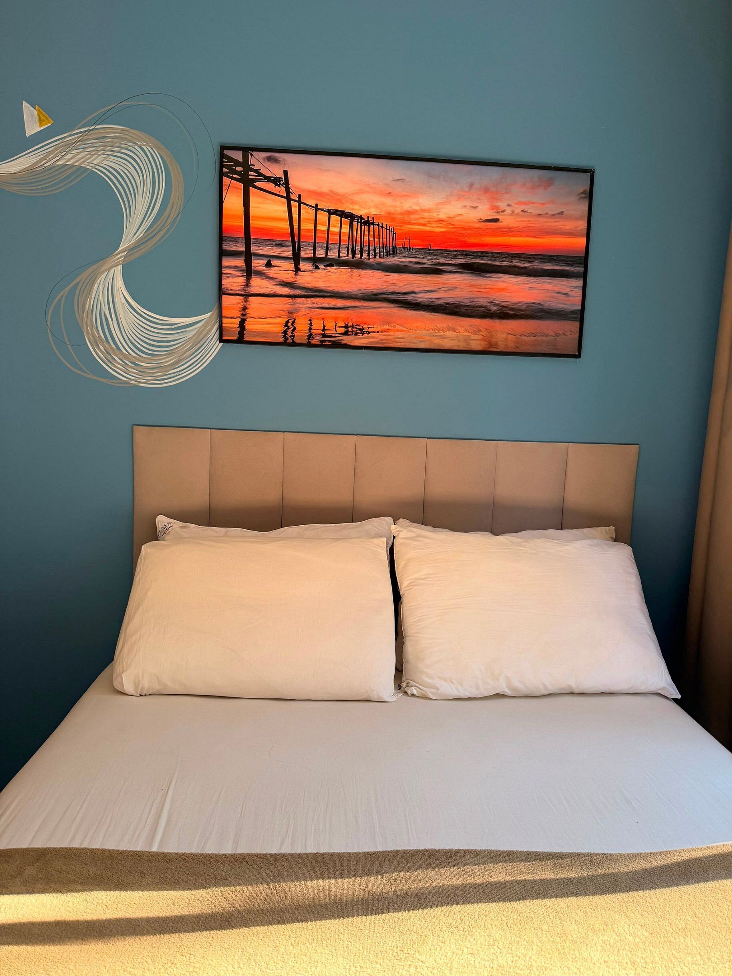 Una cama con dos almohadas, una pared con una pintura de atardecer y un adorno de pared.