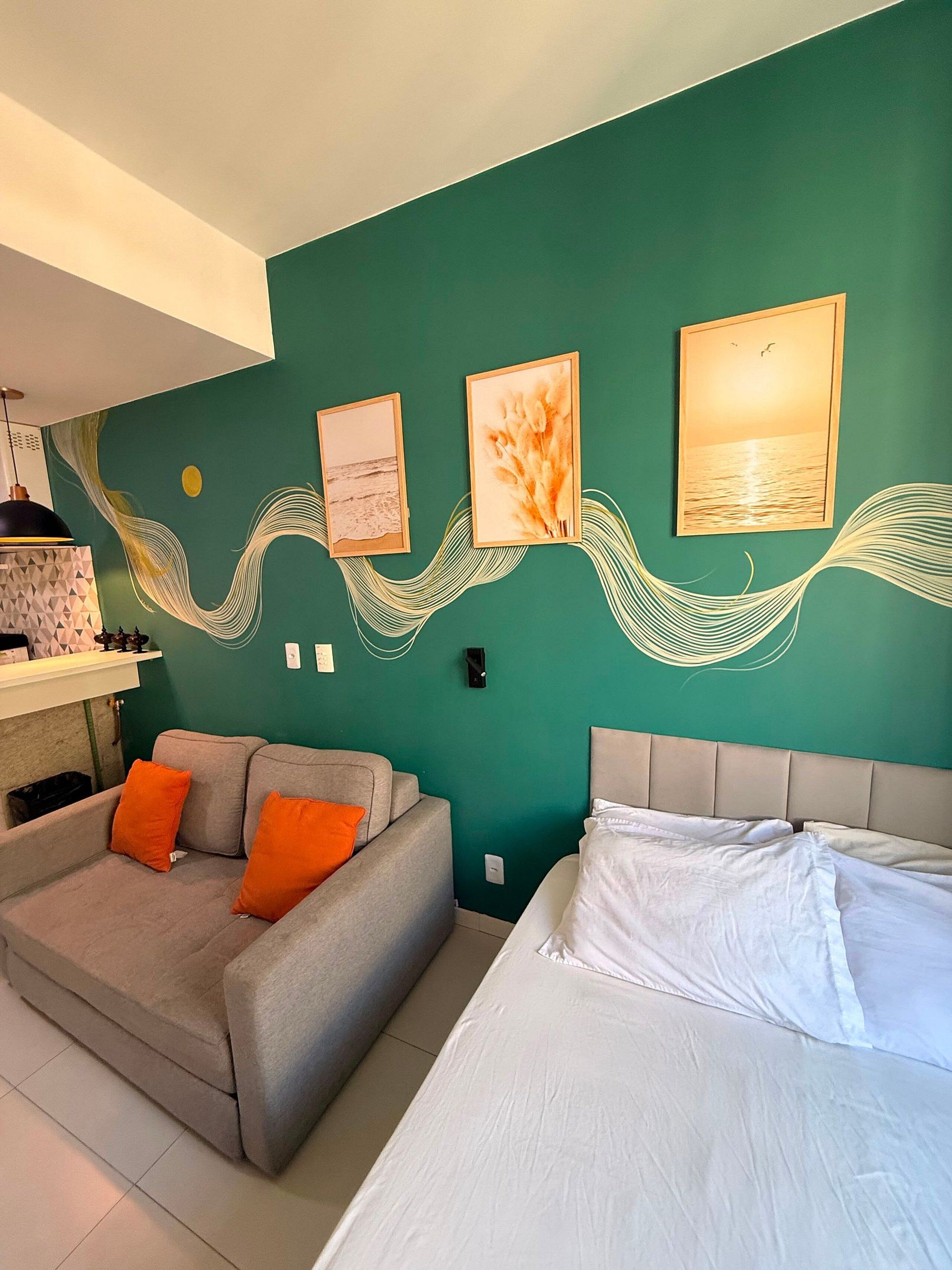 Una habitación con pared color turquesa, un sofá con cojines naranja, una cama con ropa blanca y tres cuadros enmarcados que muestran paisajes.