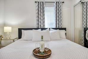 A neatly made bed with white linens, a bedside table with a lamp, and a tray with a bottle and glasses.
