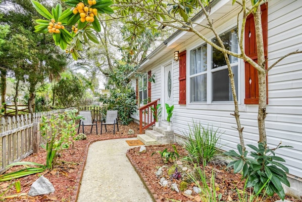 A house with a pathway leading to a porch, surrounded by various plants and a wooden fence.