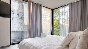A modern bedroom with a large bed, a view of trees through sheer curtains, and a concrete wall. - Kite Suite 207 (Tulum)