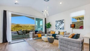 A spacious living room with a large sliding glass door leading to an outdoor patio, a grey sofa set, a coffee table, and a modern chandelier. - New Large Home-Views-Mountain, Lake & Golf (Indio)