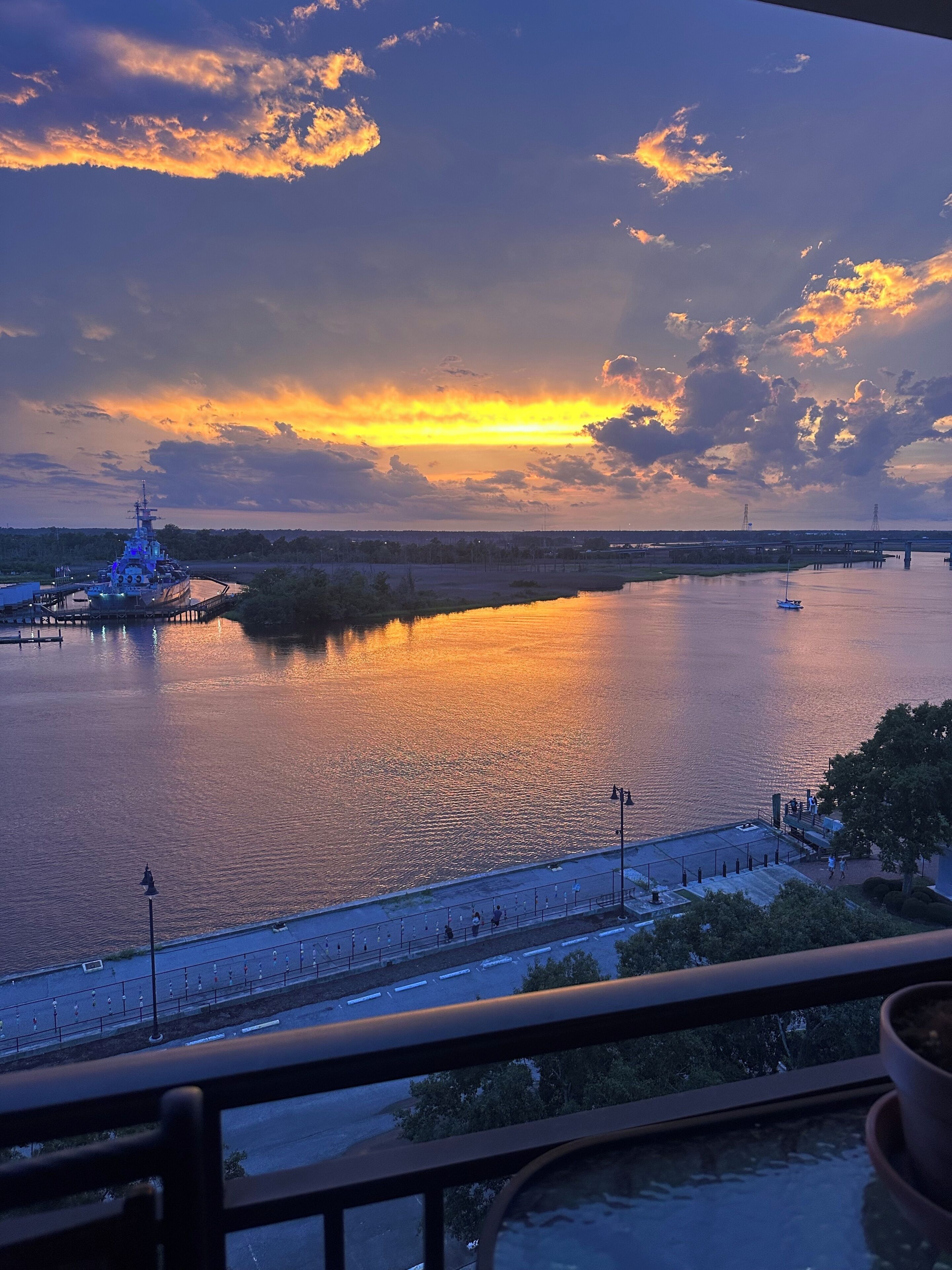 A sunset view from a balcony overlooking a river with a ship docked on the left and a pedestrian walkway in the foreground.