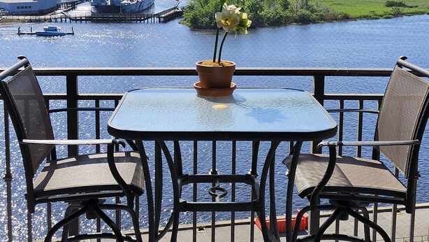 Un balcón con mesa y sillas con vista a un río, un barco atracado al fondo y una zona verde al otro lado del agua.