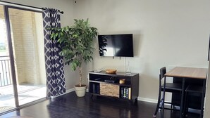 Una sala de estar moderna con piso de madera oscura, ventilador de techo, televisor de pantalla plana, mesa de comedor y una planta en maceta.