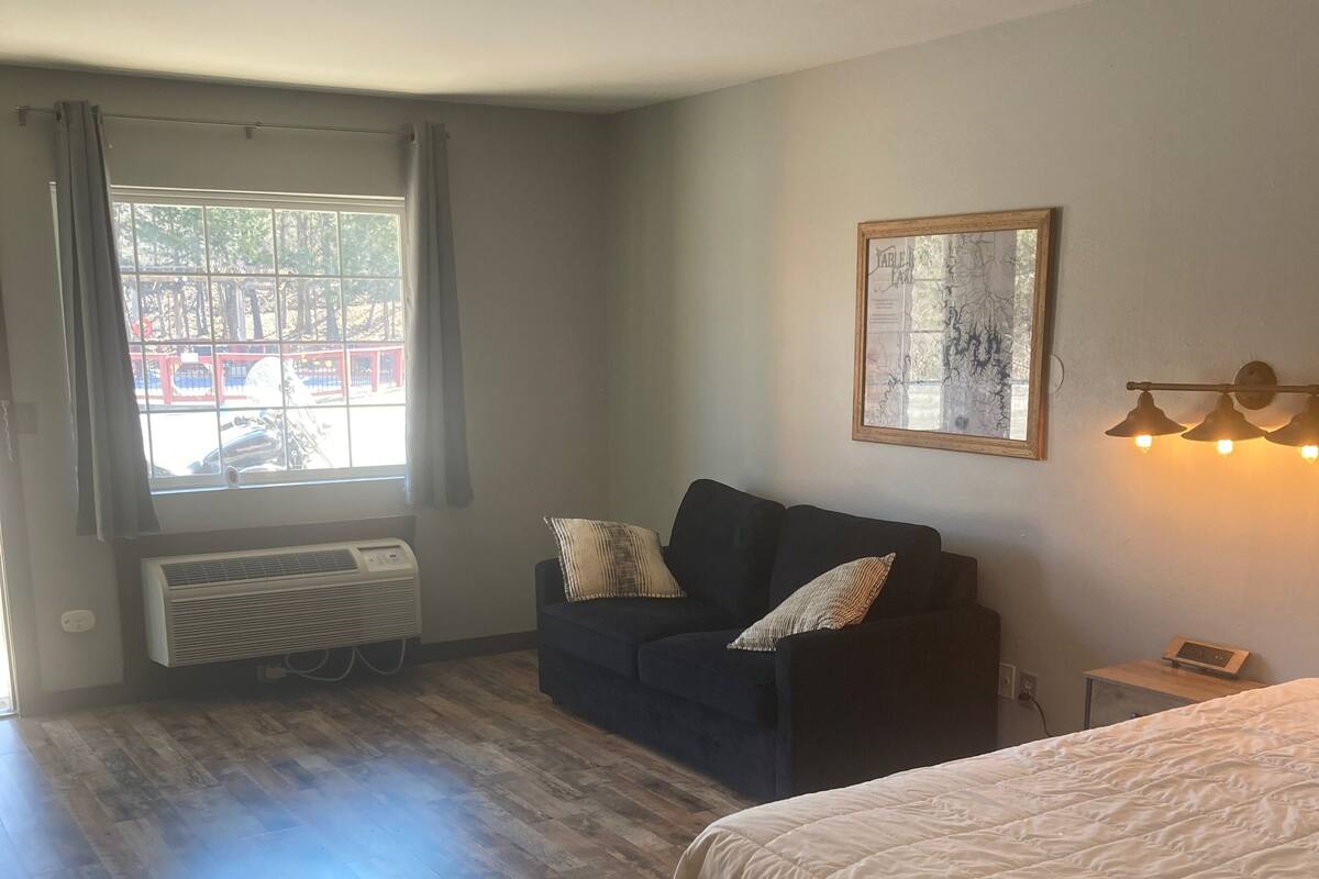 A room with a bed, a dark-colored sofa with cushions, a map on the wall, a window with curtains, and a radiator.