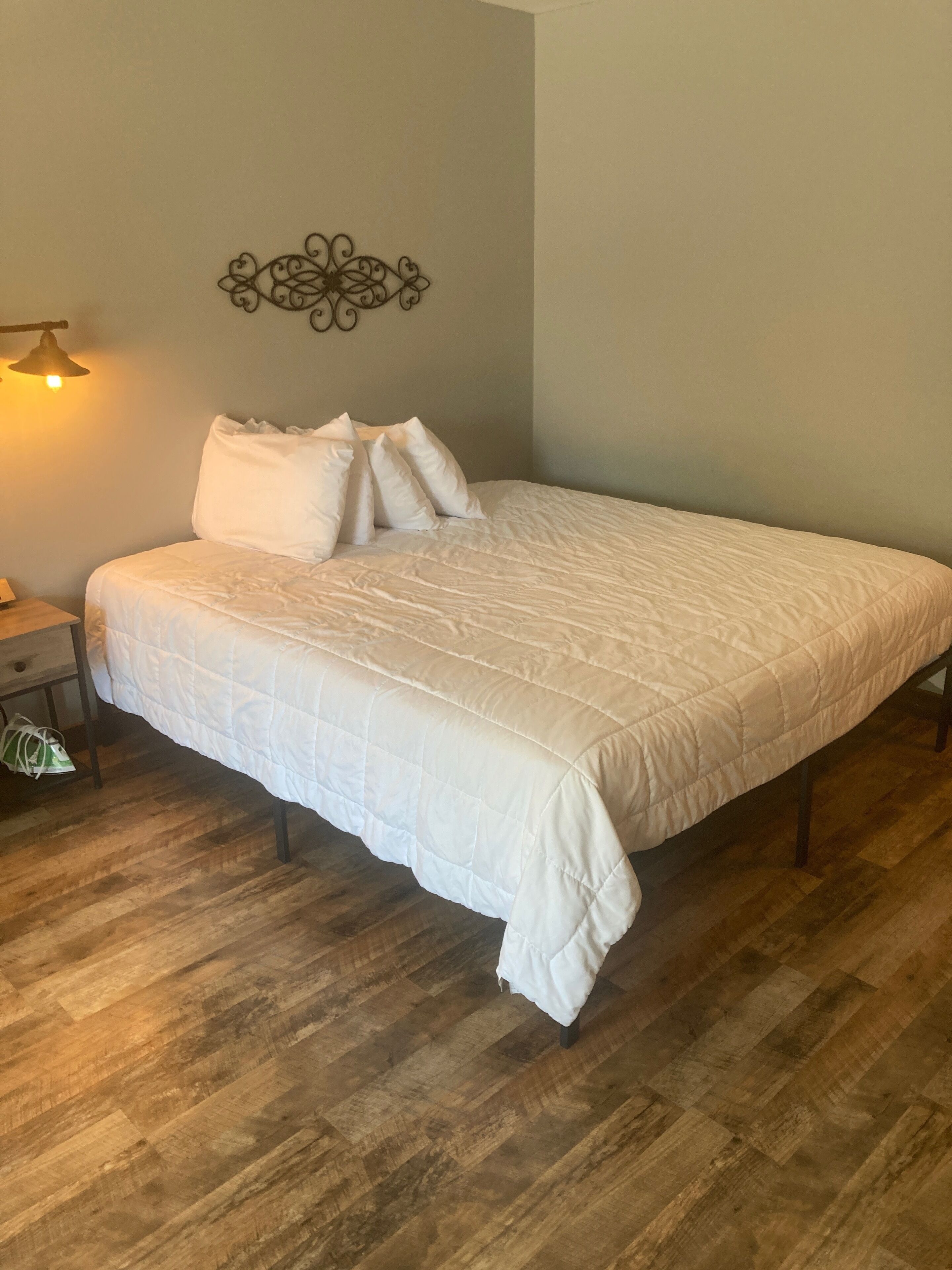 A bedroom with a large bed, wooden flooring, a wall-mounted light fixture, and a decorative wall piece.