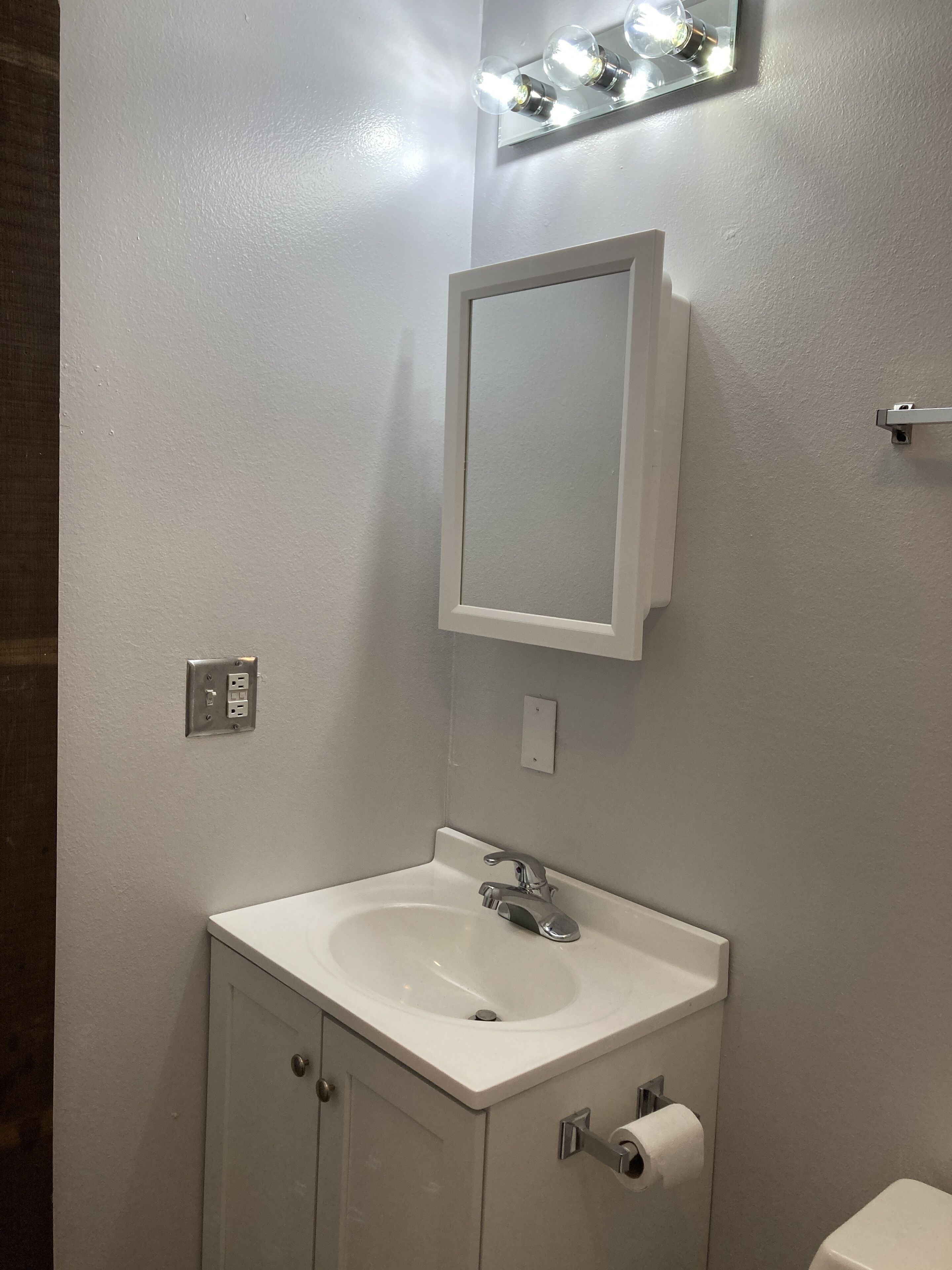 A bathroom with a white sink, mirror, and vanity. There are lights above the mirror and a towel rack to the right.