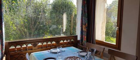 A dining table set with plates, glasses, and a cake, overlooking a garden with trees and a stone structure.