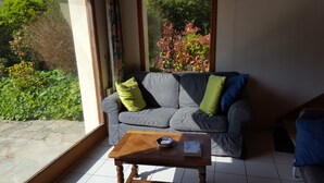 A room with a grey sofa, two cushions, a wooden coffee table, and a window overlooking greenery. - Joli Cottage en Pierres Debouts Dans un Beau Jardin Fleuri Proche Plage (Nevez)
